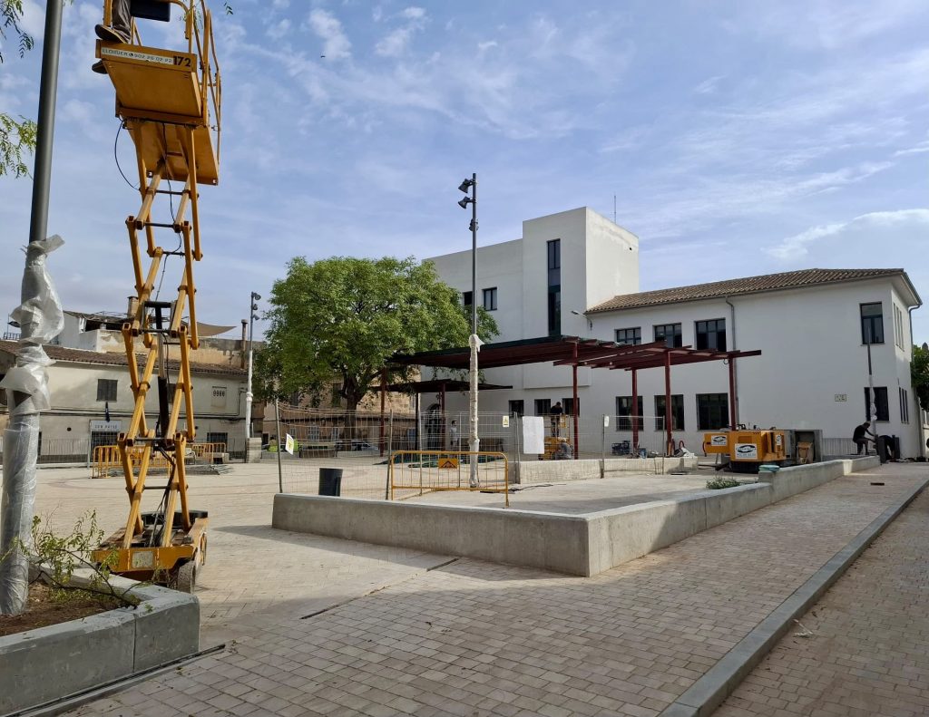 Imatge il·lustrativa d'una plaça en obres amb arbres joves, paviment de cautxú i pèrgoles pendents d'acabar.