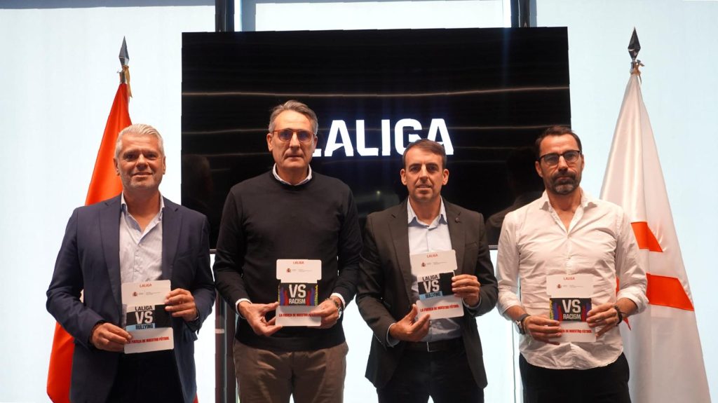 Braçalet esportiu de color viu amb missatge contra el bullying i el racisme, col·locat sobre la gespa d'un camp de futbol, amb llum natural de dia.